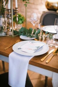 In questa foto un posto a tavola allestito con piatto bianco in porcellana e il suo menù, bicchieri di cristallo trasparente e posate silver